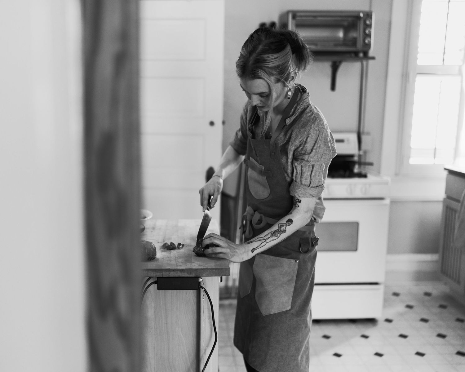 Woman in apron using power sander on wood in a kitchen workshop, black and white photography.