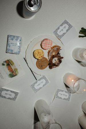 Overhead view of a white wedding reception table with assorted cookies, place cards, candles, and decorative matchboxes.