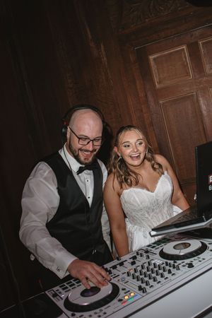 Laughing bride in white gown and groom in black vest enjoy DJing together at their wedding reception.