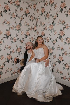 Bride and groom sitting against floral wallpaper, holding drinks in a candid wedding portrait.