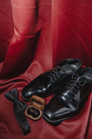 Black patent leather dress shoes with bow tie and wooden ring box on red satin fabric for wedding.