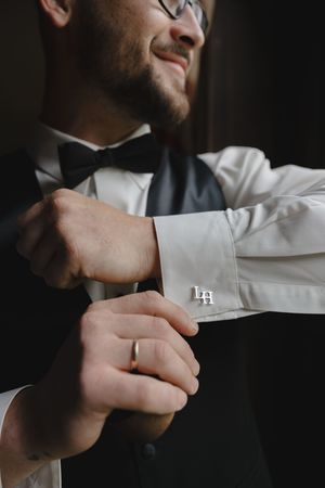 Smiling groom adjusting monogrammed cufflinks on white dress shirt wearing black bow tie and wedding ring.