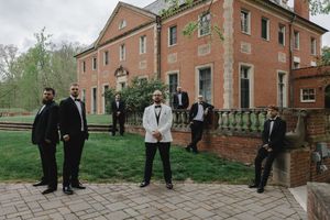 Groom and groomsmen in black tuxedos pose outside a historic red brick mansion on a wedding day.
