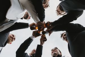 Men in suits toasting with whiskey glasses in a circle, shot from below at a wedding celebration or formal event.