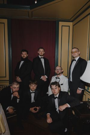 Seven groomsmen in black tuxedos pose elegantly in a dimly lit vintage room with burgundy and gold decor.