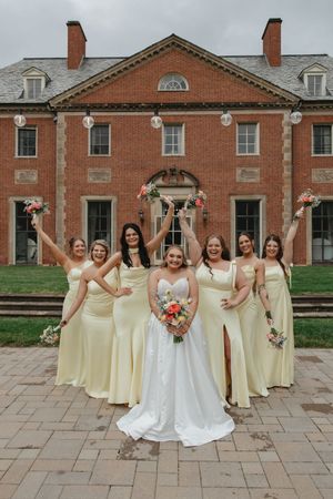 Bridesmaids in yellow dresses raise bouquets celebrating with bride in white gown outside brick mansion.