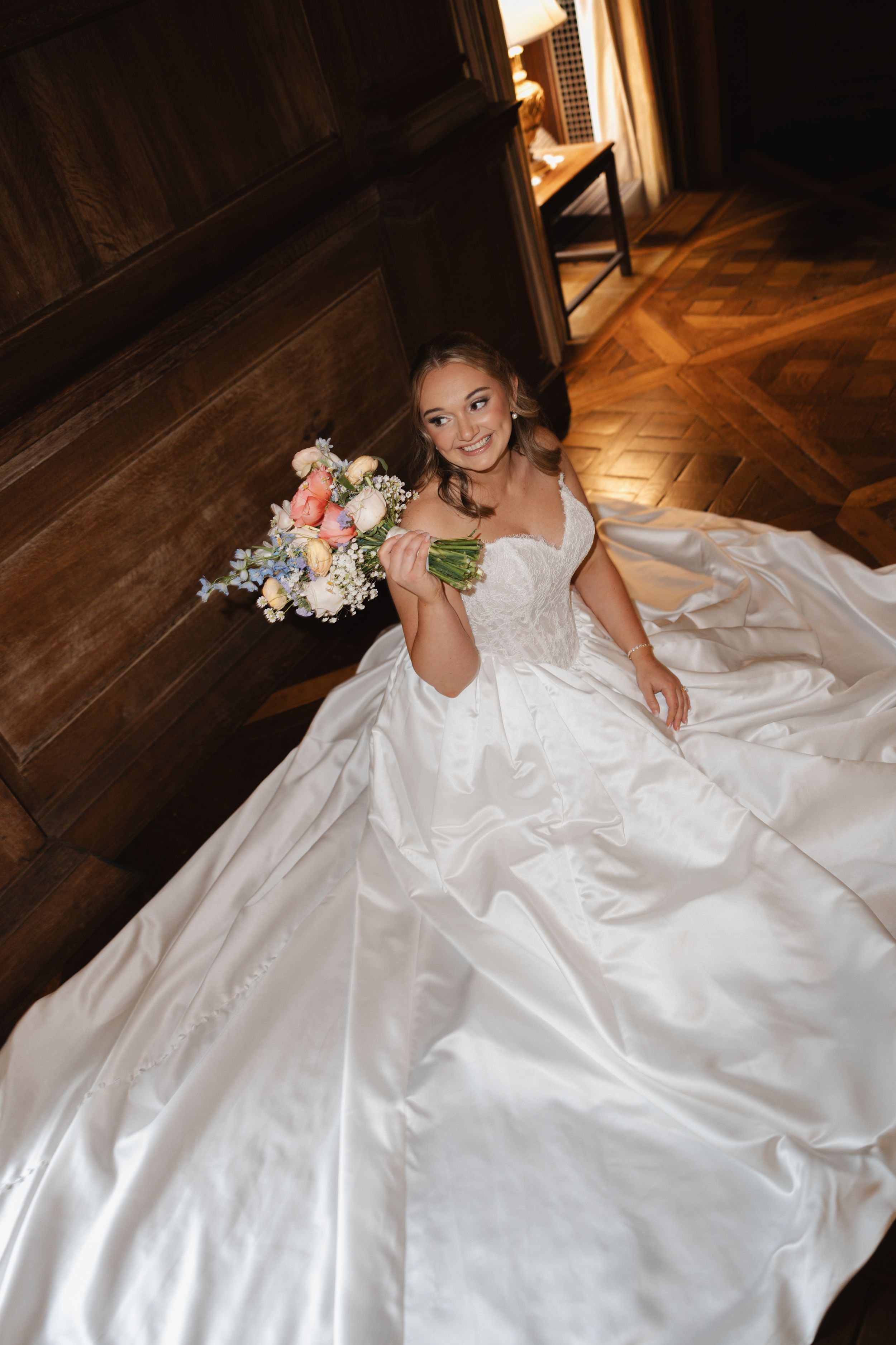 Bride in a voluminous white ball gown sits on wooden floor holding a colorful spring bouquet, smiling up at camera.