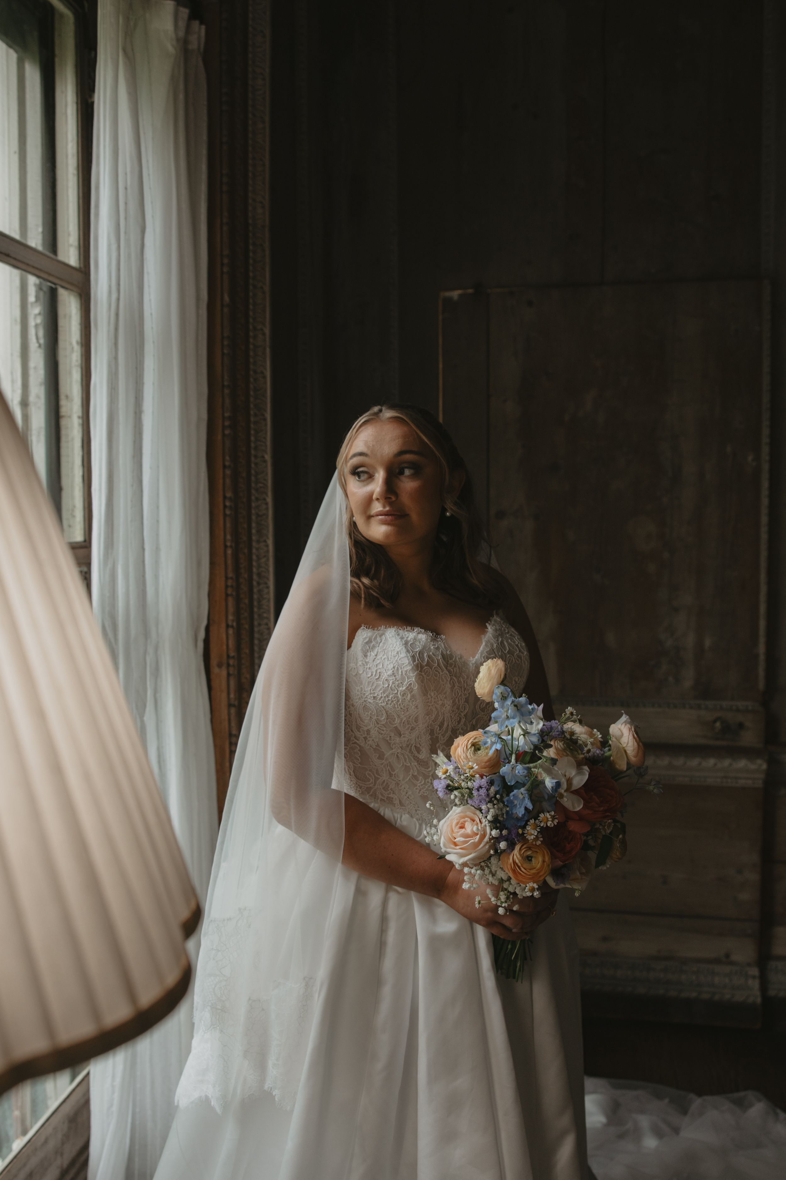 Elegant bride in lace wedding gown and veil stands by window holding a pastel blue and peach floral bouquet.