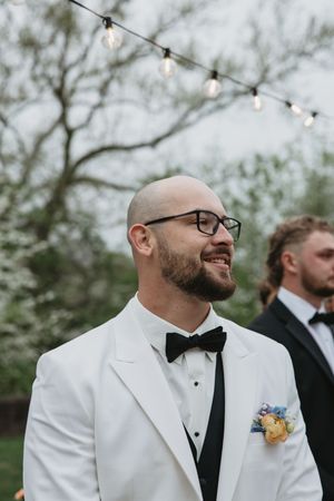 Smiling bald groom in white tuxedo jacket and black bow tie stands outdoors with string lights behind him.