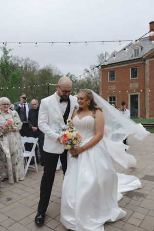 Bride and groom walking down outdoor wedding aisle, bride holding colorful bouquet with veil flowing in wind.