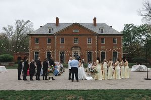 Outdoor wedding ceremony at historic brick mansion with string lights and bridesmaids in yellow dresses.