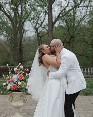 Bride and groom sharing first kiss at outdoor wedding ceremony surrounded by colorful floral arrangements.