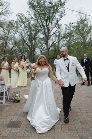 Bride in white ballgown and groom in white tuxedo jacket walk hand-in-hand after outdoor wedding ceremony.
