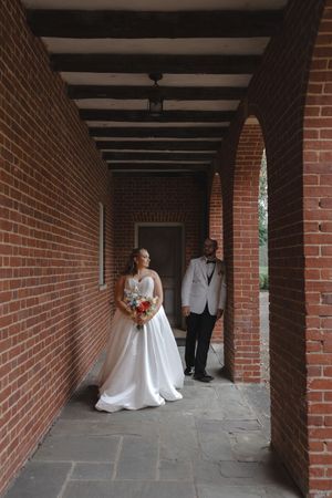 Bride in white ball gown and groom in white tuxedo standing in a brick archway corridor at their wedding venue.