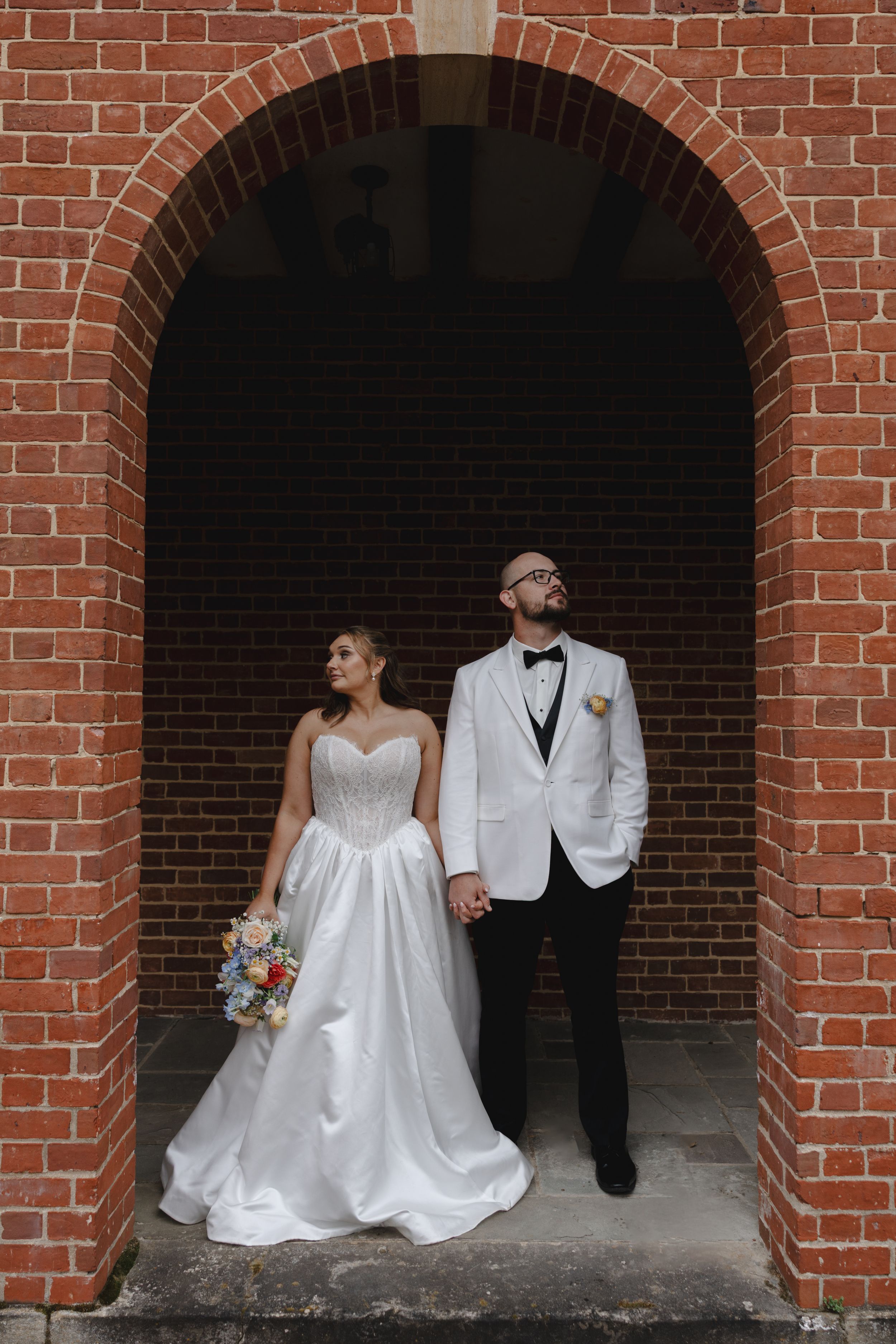 Bride and groom pose romantically under brick archway, bride holding colorful bouquet.