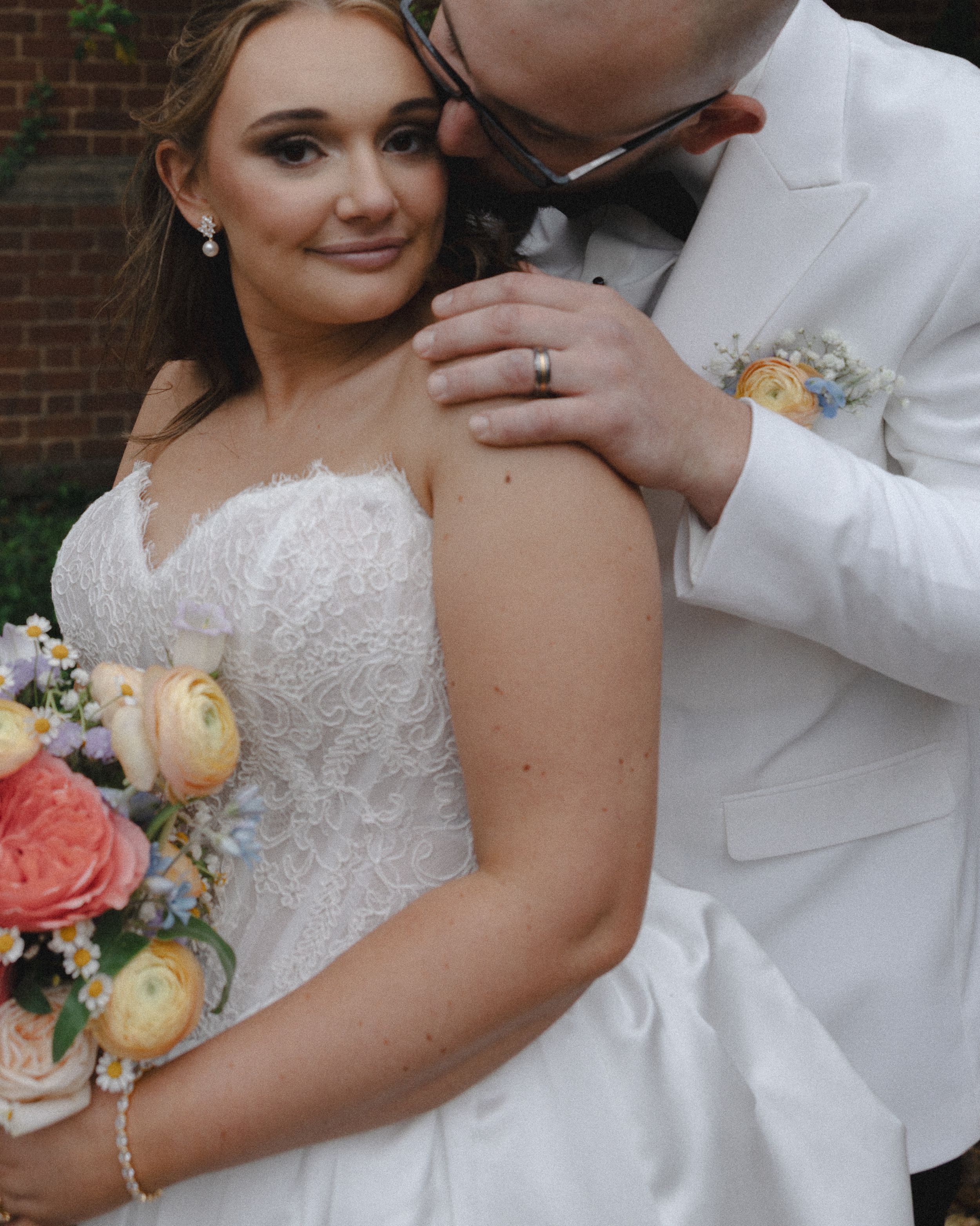 Bride in strapless lace gown holds colorful bouquet as groom in white suit tenderly kisses her shoulder.