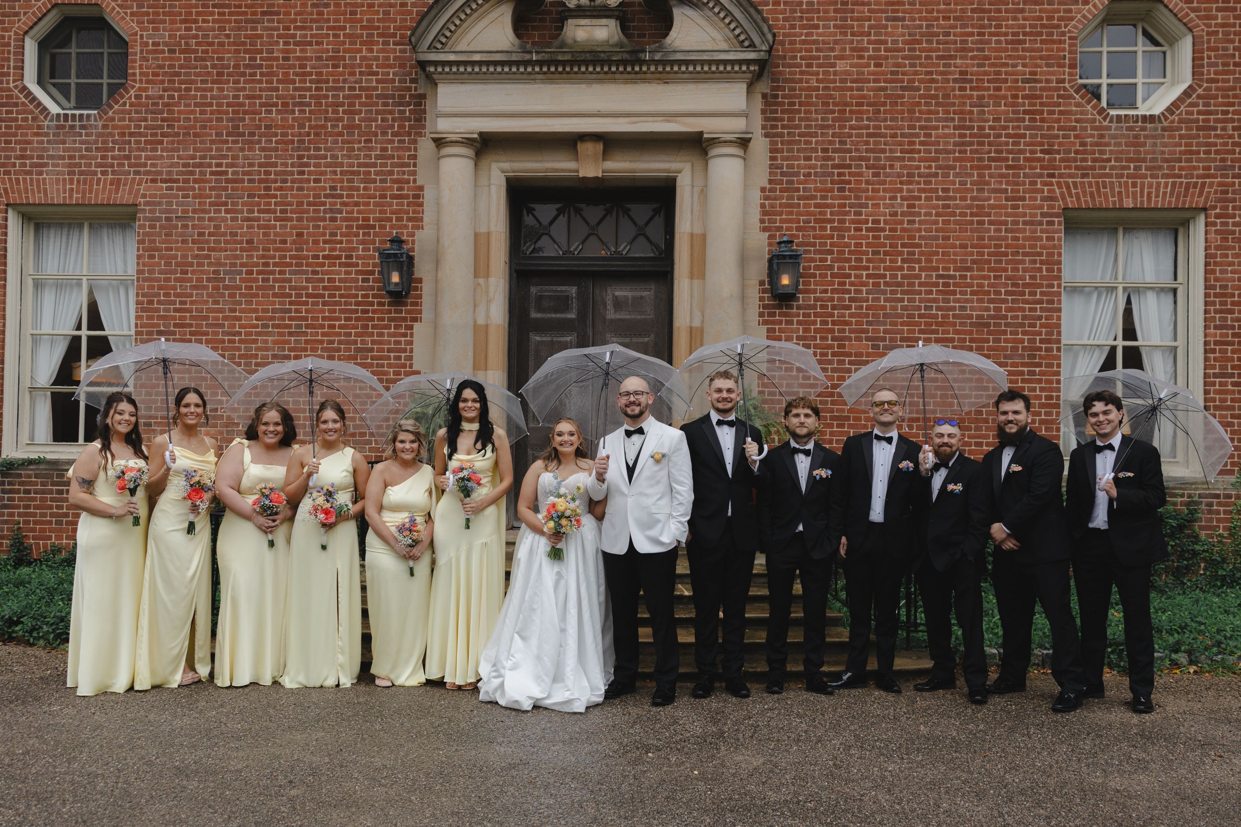 Wedding party poses in front of historic brick venue, bridesmaids in yellow gowns and groomsmen in black tuxedos.