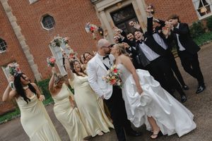 Bride and groom share a kiss surrounded by cheering wedding party outside historic brick building.