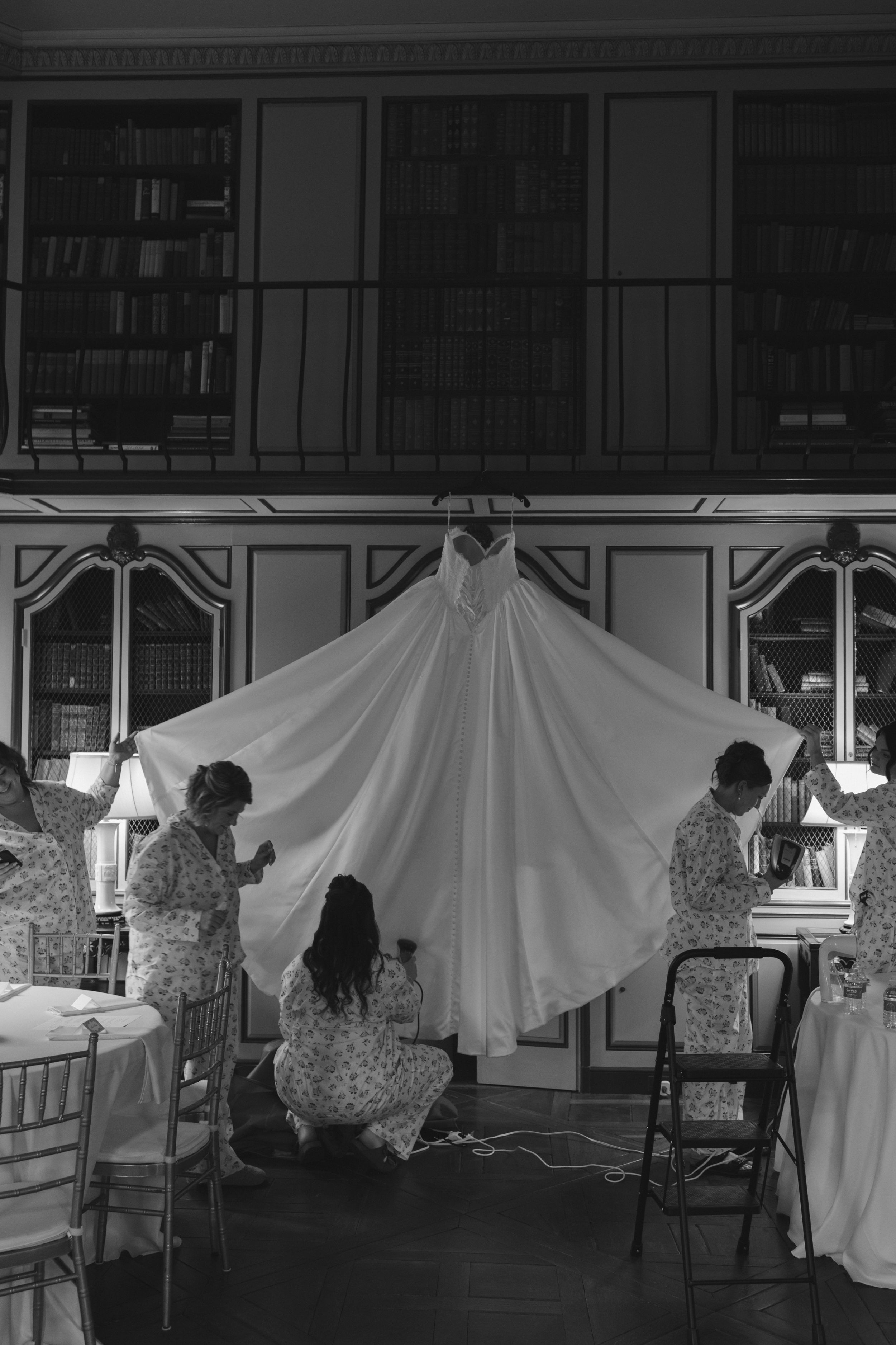 Black and white photo of bridesmaids arranging a wedding dress in an elegant library setting.