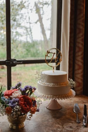 Two-tier white wedding cake with gold monogram topper on pink stand beside colorful floral arrangement near rustic window.