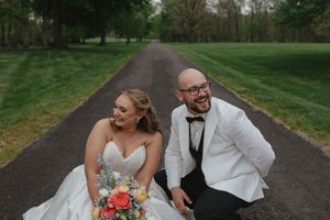 Bride and groom laughing together while sitting on a tree-lined driveway, bride holding colorful bouquet.