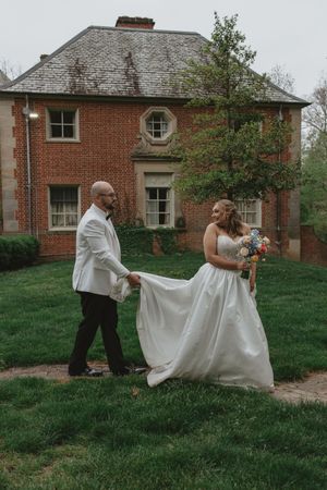 Groom in white jacket helps bride carry her white ballgown train across green lawn near brick estate.