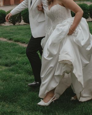 Close-up of bride in white heels and ballgown walking with groom across lush green grass.