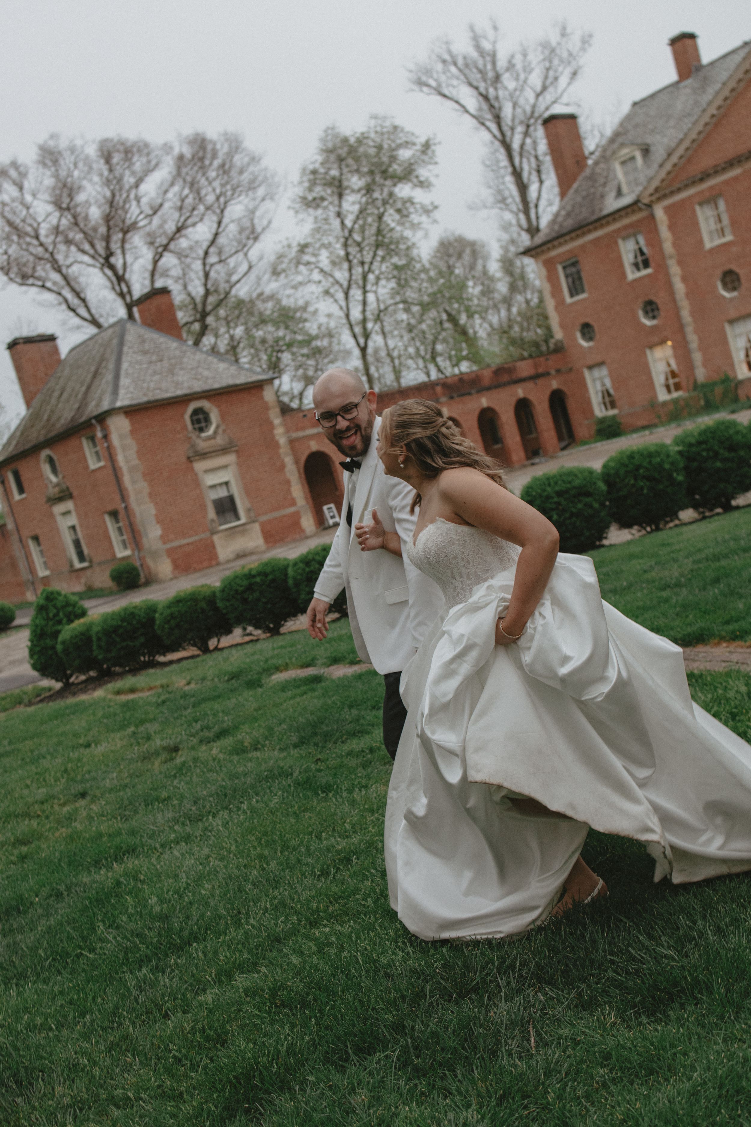 Bride and groom run playfully across green lawn outside a historic red brick estate on their wedding day.
