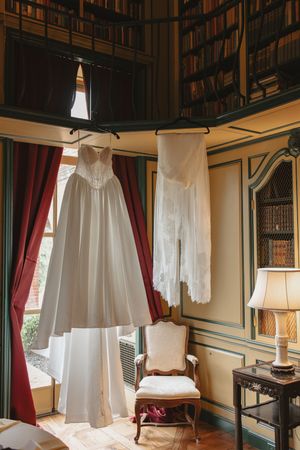 Elegant white wedding dress and veil hanging in a vintage library room with red curtains and antique chair.