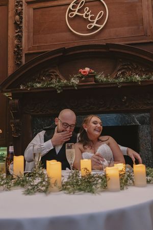 Bride and groom laughing at sweetheart table with candles and floral decor before ornate wood fireplace.