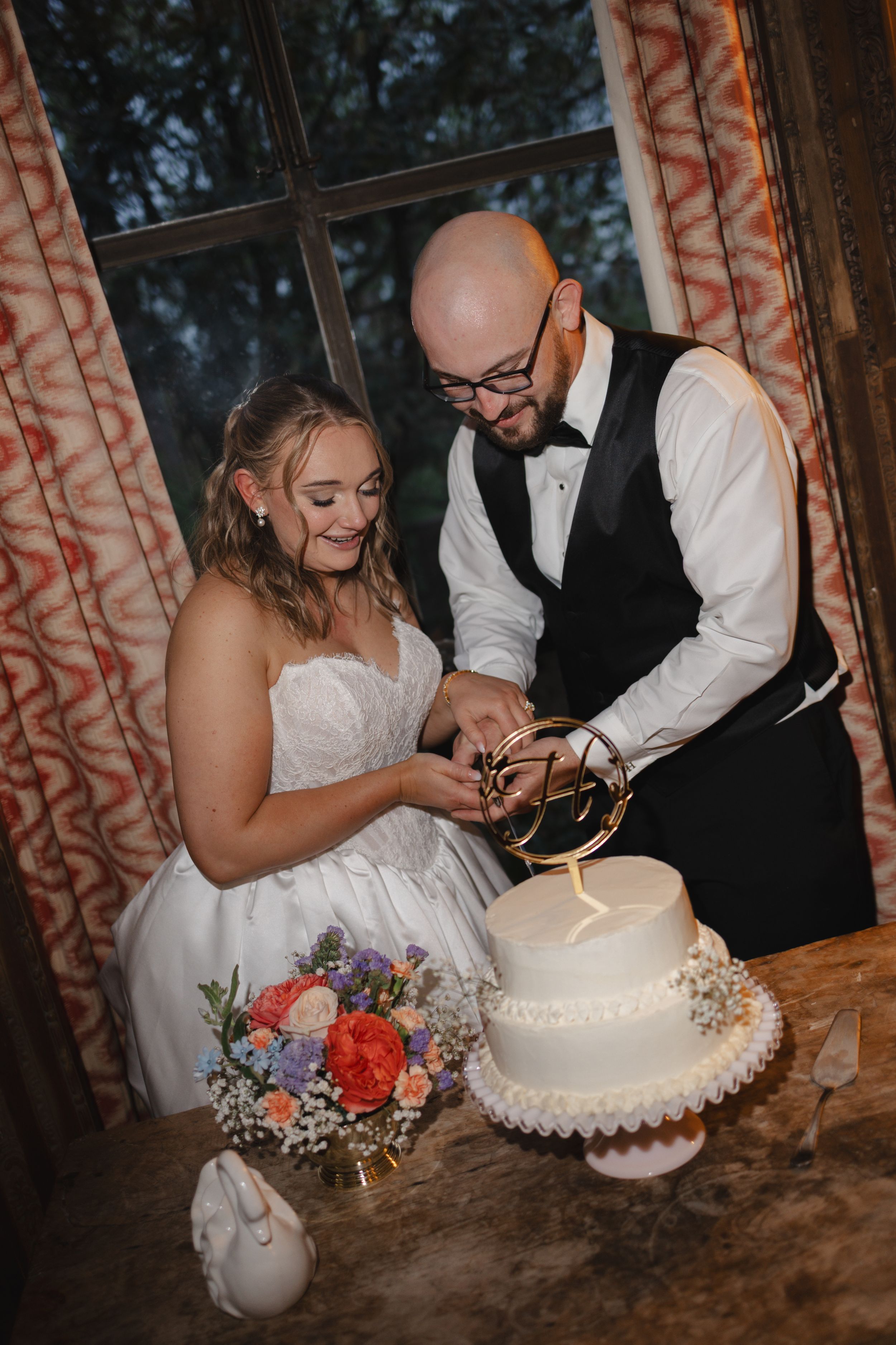 Smiling bride and bald groom cut their white wedding cake adorned with a gold topper at their reception.