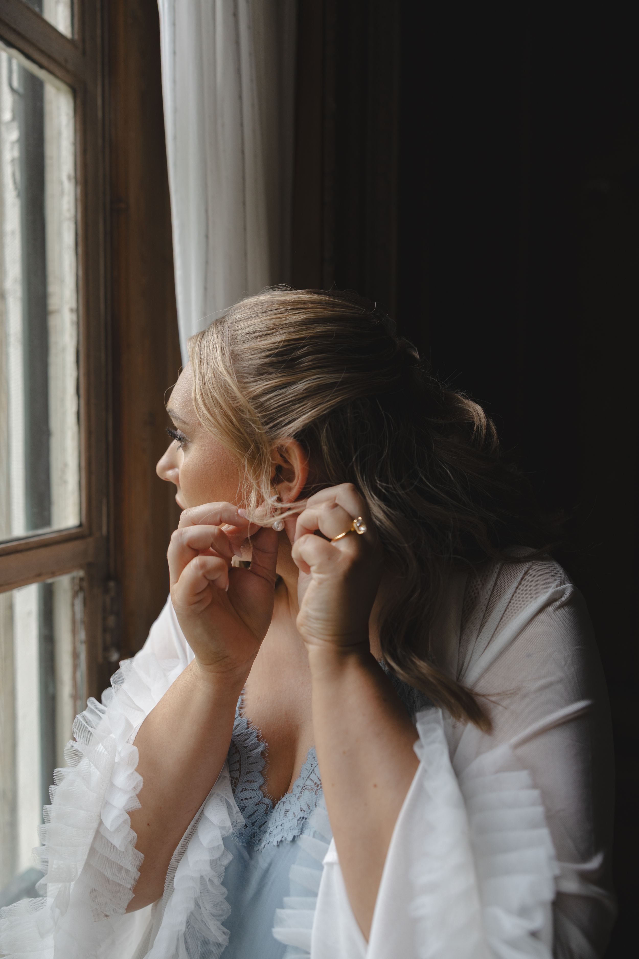 Blonde bride in white robe putting on earring while gazing out dark wooden window during wedding preparations.