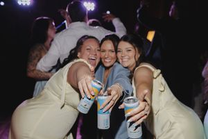 Three women smiling and holding canned drinks together on a dark dance floor at a wedding reception.