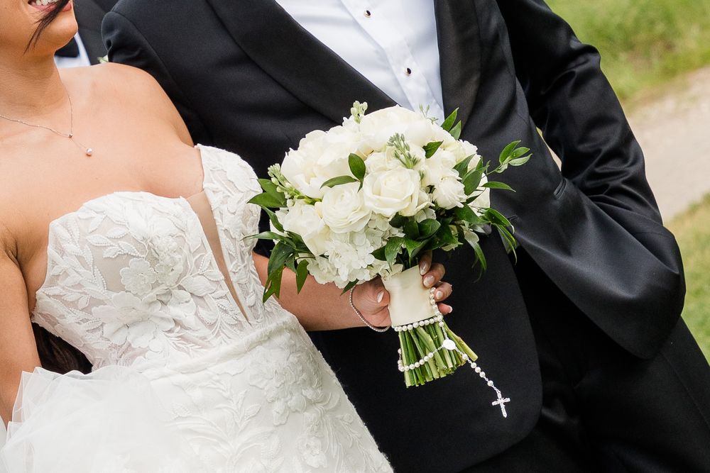 Bride and groom holding a white rose and greenery wedding bouquet with a rosary, wearing classic black tuxedo and white lace gown.