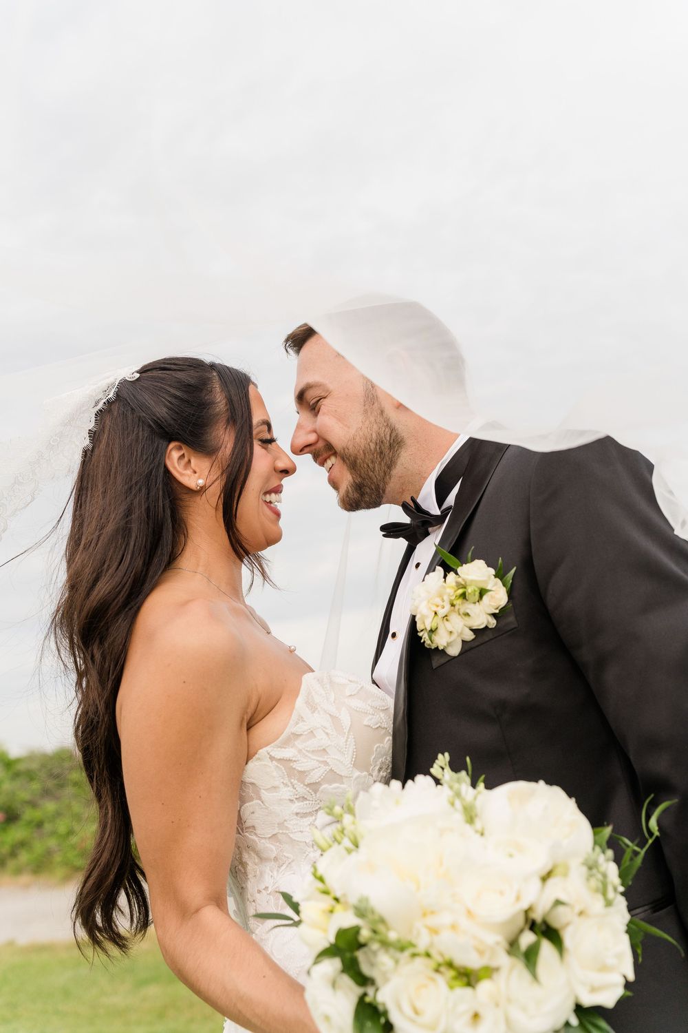 A bride and groom share a romantic nose-to-nose moment, she in a strapless gown holding white roses, he in a black tuxedo.