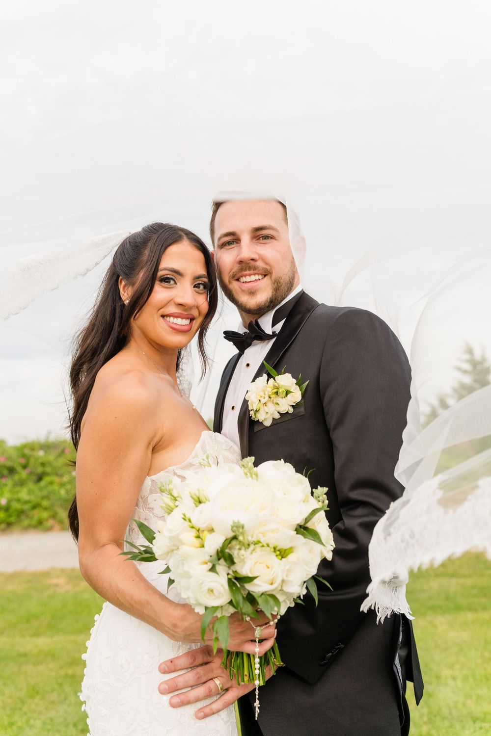Bride and groom posing outdoors on wedding day, bride holding white floral bouquet with veil flowing in breeze.