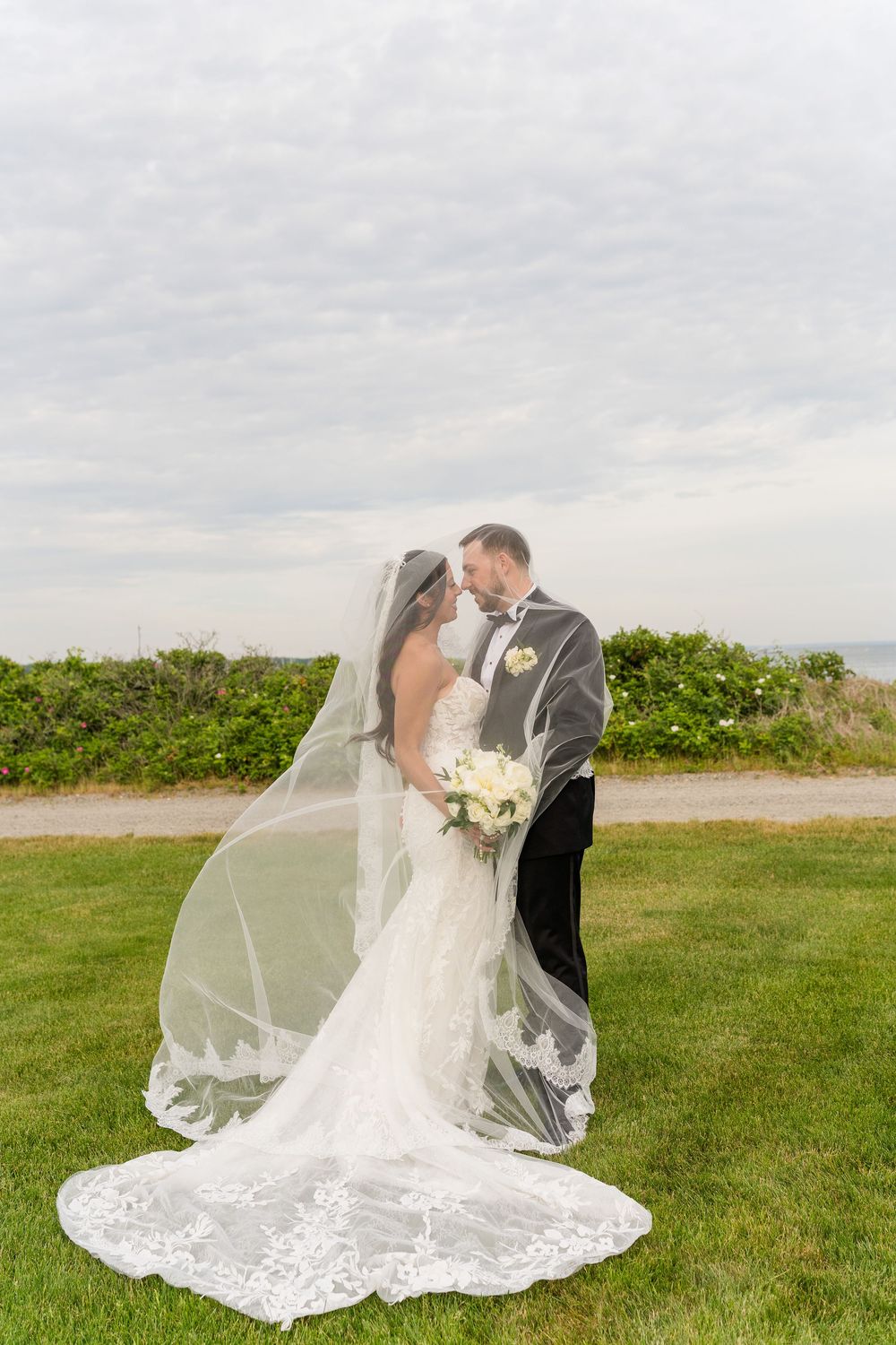 Bride and groom share a romantic moment outdoors, her lace veil flowing in the wind against a cloudy sky.