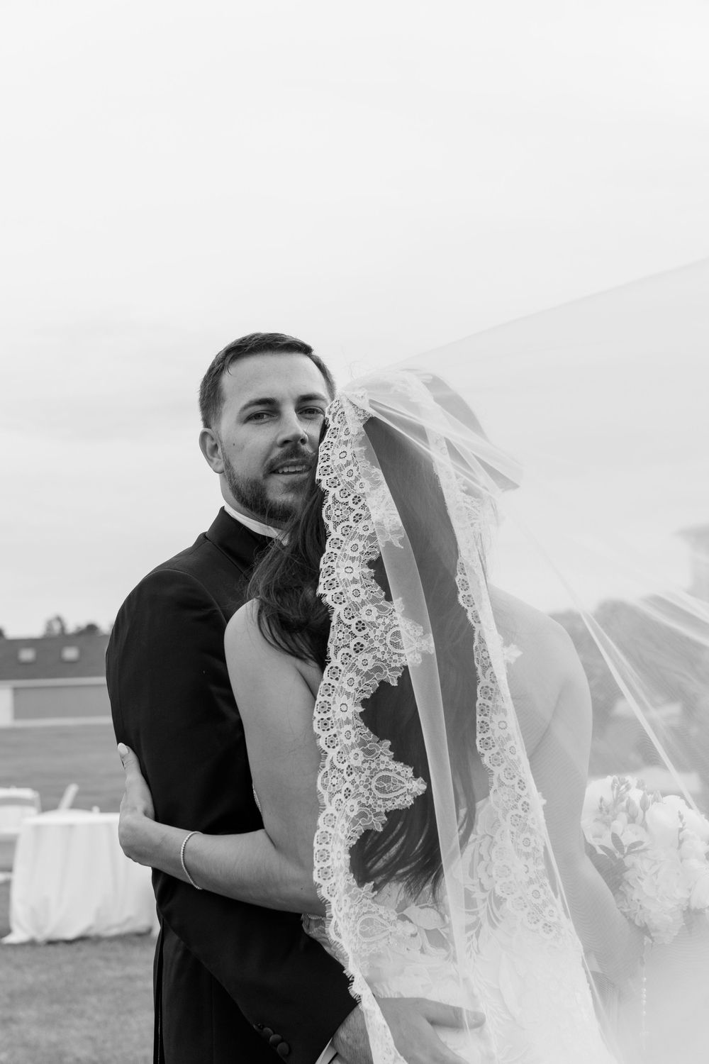 Black and white wedding photo of groom embracing bride in lace veil outdoors.