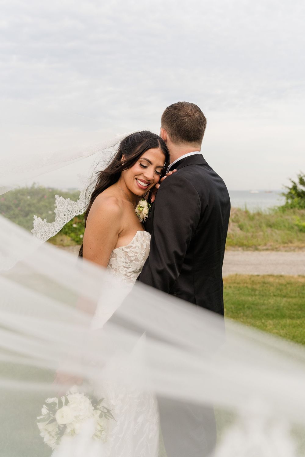 Bride smiles at groom as veil blows in wind outdoors.