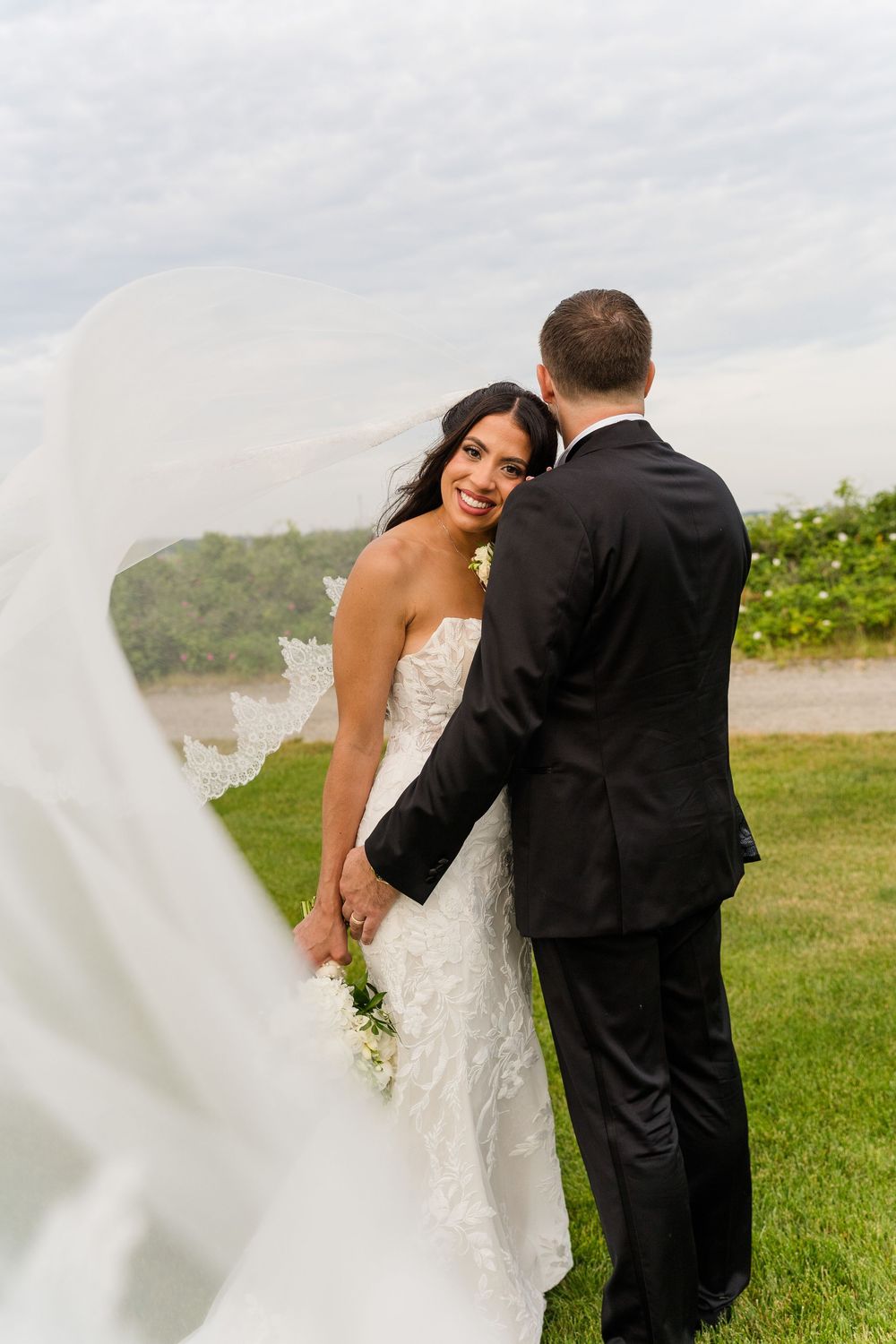 Bride smiling over shoulder at groom while veil billows in wind during outdoor wedding portrait on green lawn.