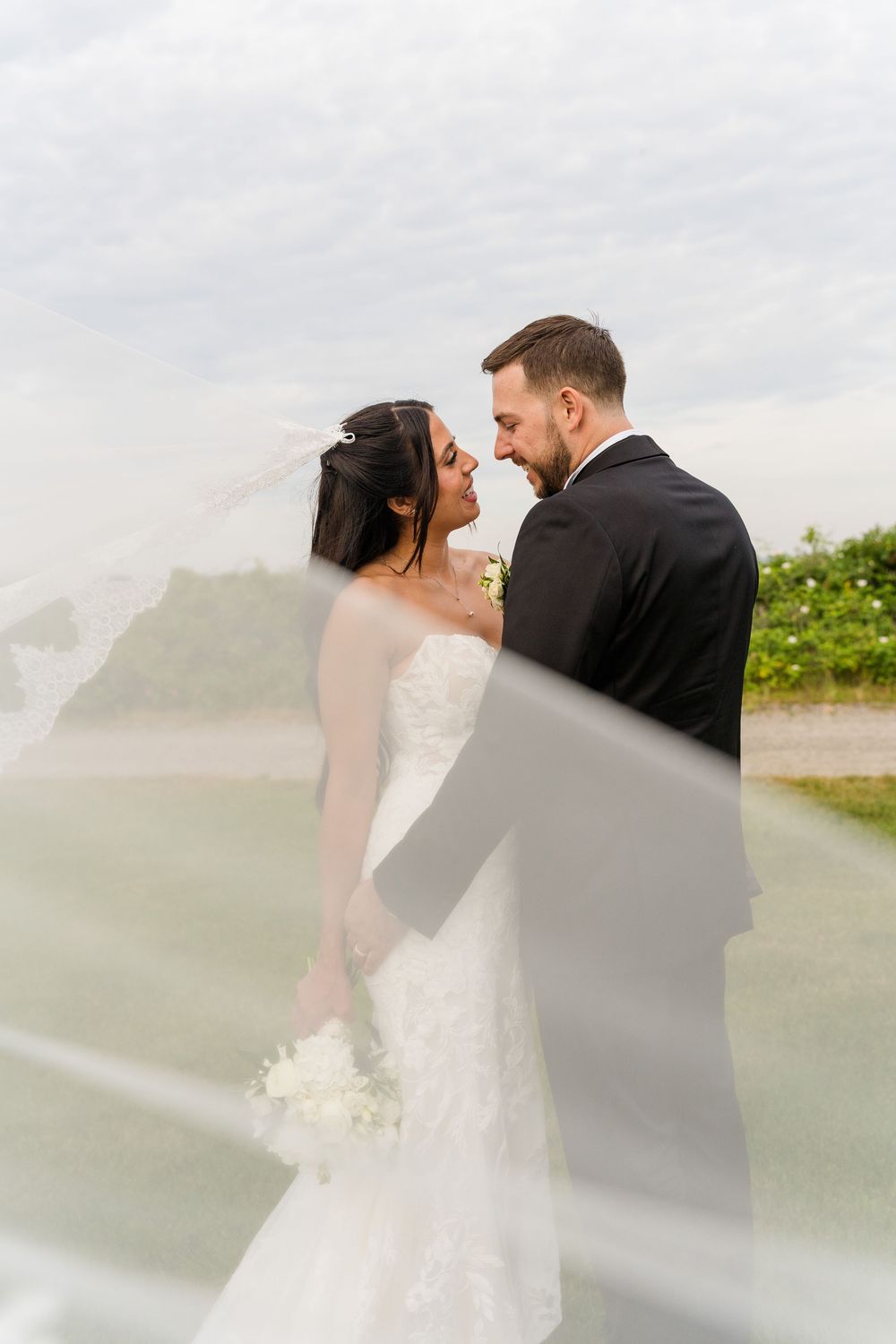 Bride and groom share a romantic moment as her veil billows in the wind at an outdoor wedding.