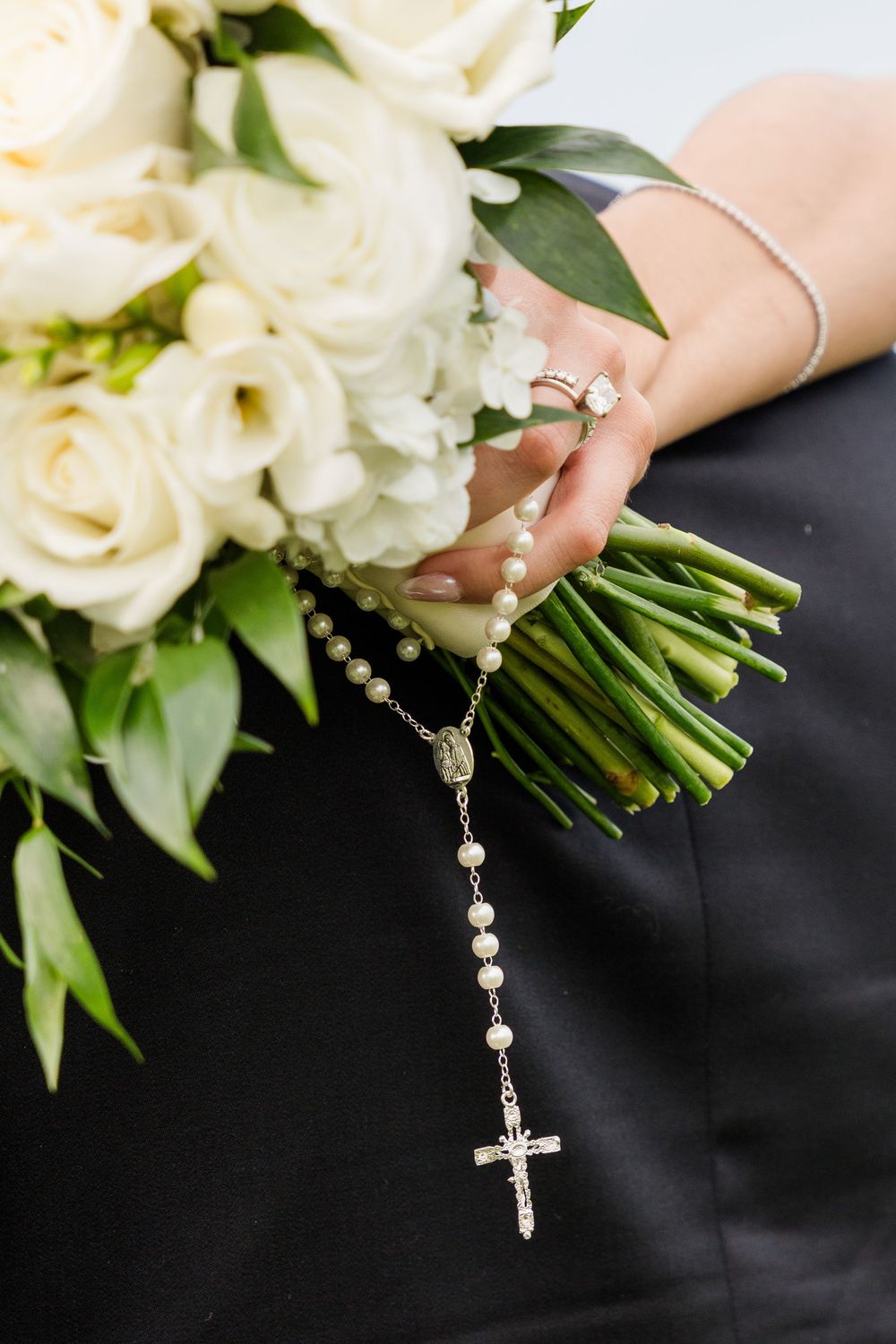 Bride holding white rose bouquet with pearl rosary and diamond ring on wedding day.
