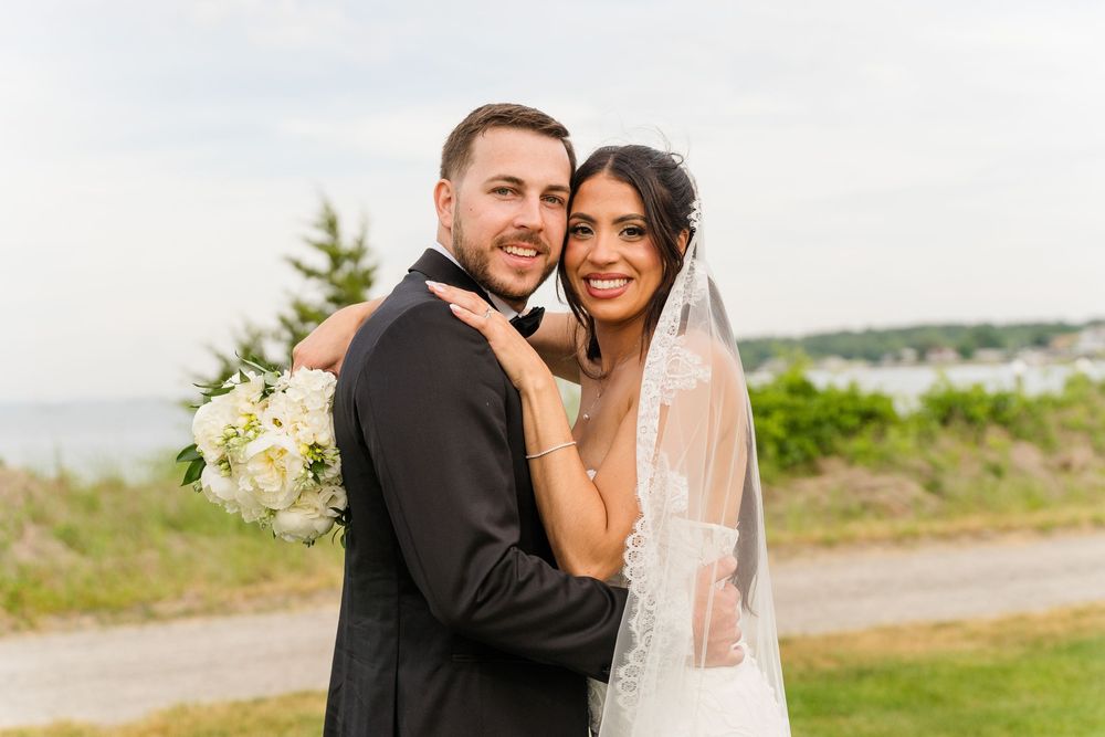 Bride and groom embracing outdoors, bride holding white bouquet and wearing lace veil, smiling on their wedding day.