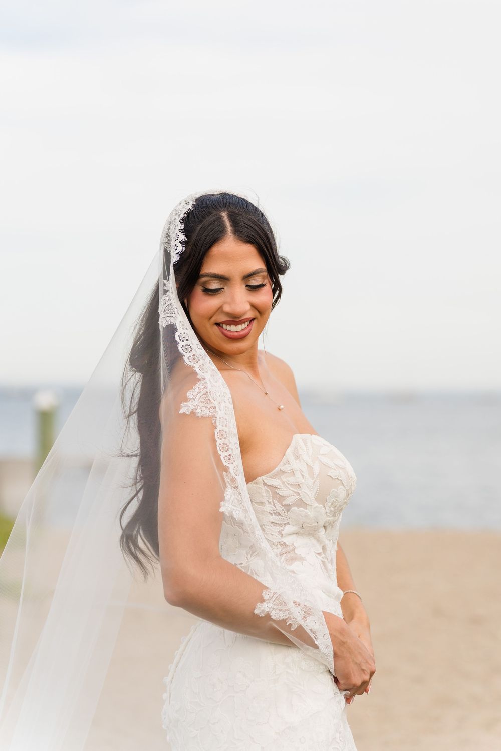 Smiling bride in lace wedding dress and veil poses on beach with ocean backdrop.