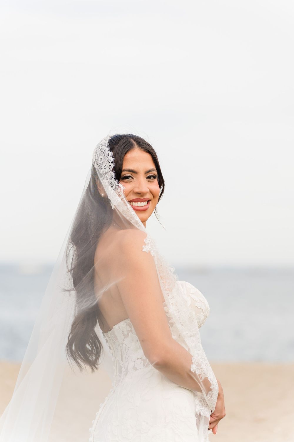 Smiling bride in lace wedding gown and veil poses on sandy beach with ocean backdrop.