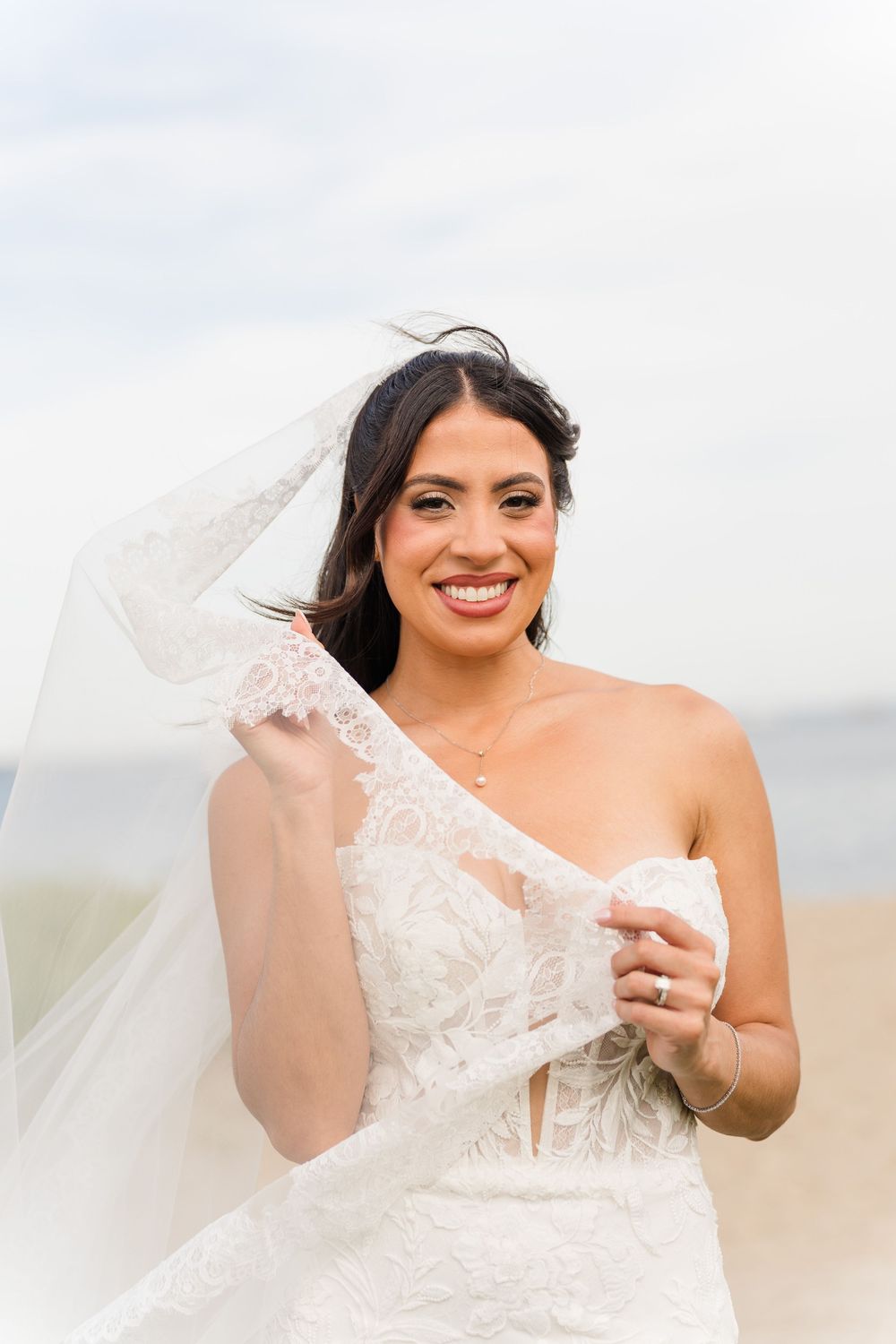 Smiling bride in lace wedding gown holds veil at beach.