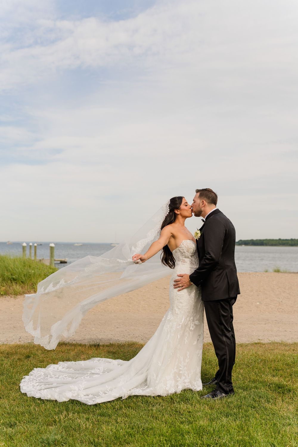 Bride and groom share a romantic kiss outdoors by the waterfront, with her lace gown and veil flowing in the wind.
