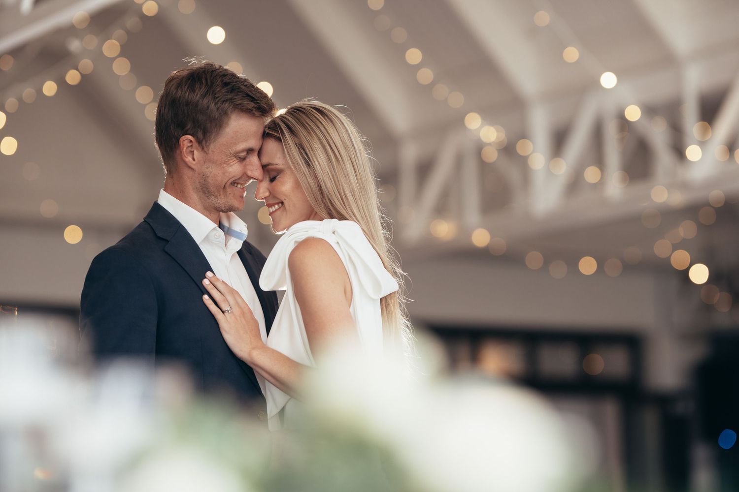 Bride and groom share a joyful forehead-to-forehead moment under sparkling fairy lights at wedding reception.