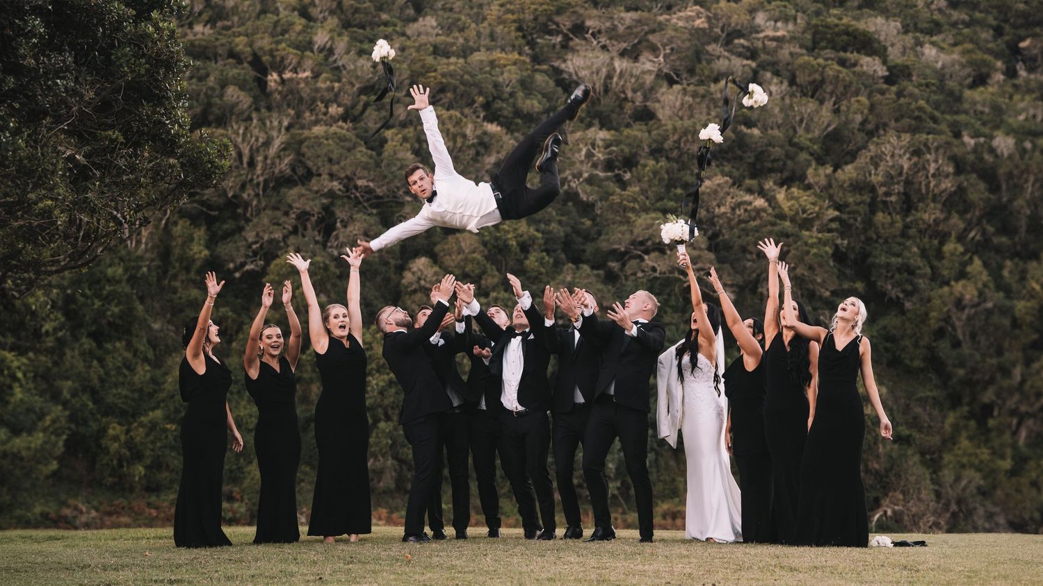 Wedding party in black formal wear tosses a groom into the air outdoors with hills behind them.