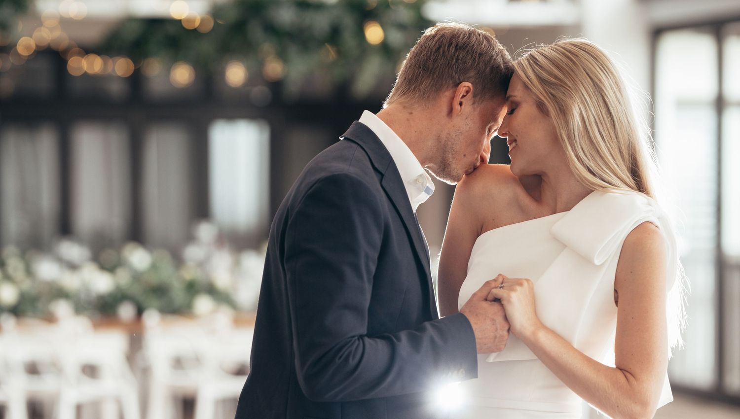 Elegant blonde bride and groom lean in for a kiss in a beautifully decorated wedding venue.